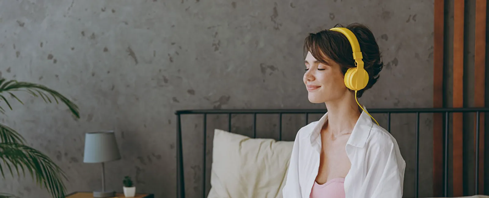 Young woman in pajamas meditating on bed with yellow headphones in cozy bedroom with soft lighting.