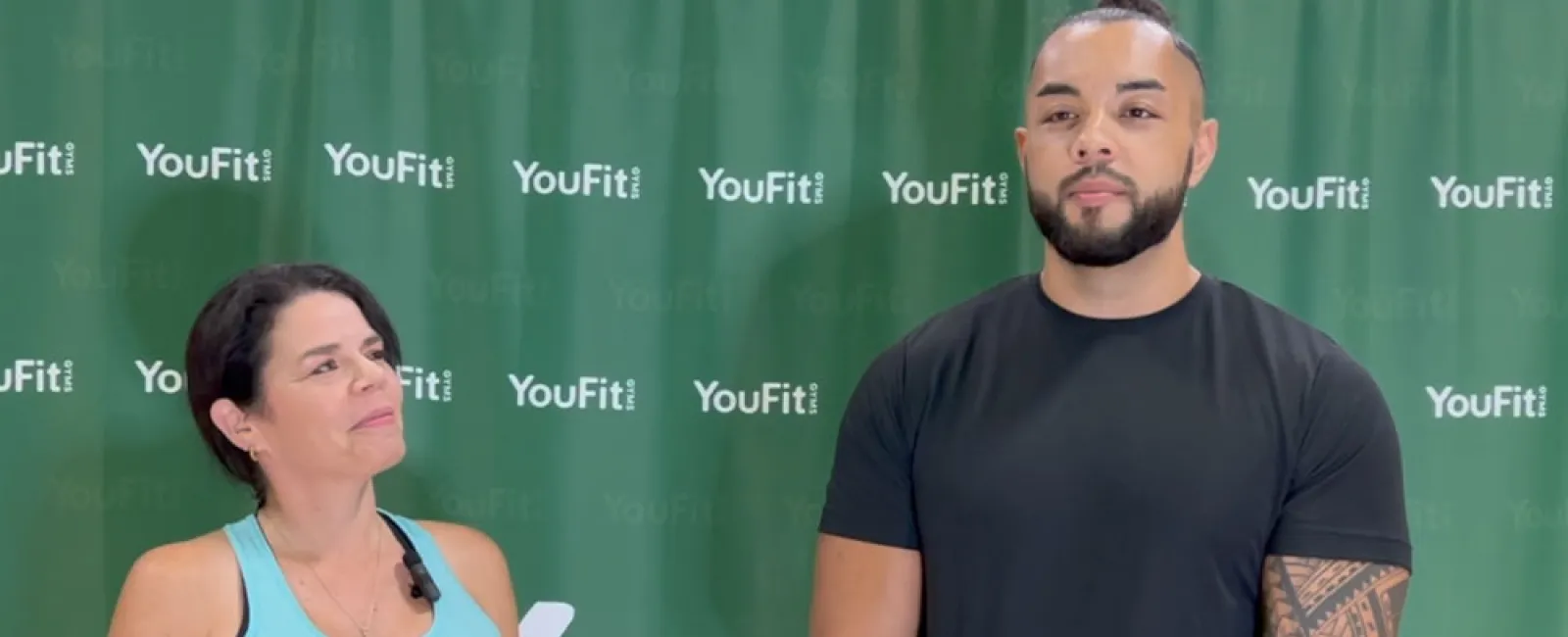 Fitness trainer and participant posing in front of YouFit gym backdrop wearing workout attire.