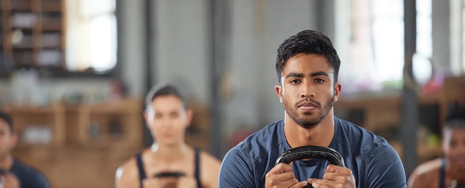 Man in blue shirt holding kettlebell, exercising with group in gym during strength training session.