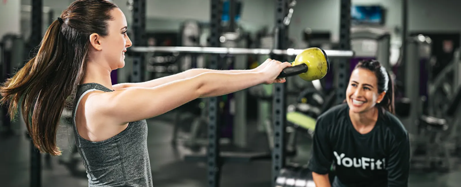 Woman lifting a kettlebell while a trainer encourages her in a gym setting with fitness equipment.