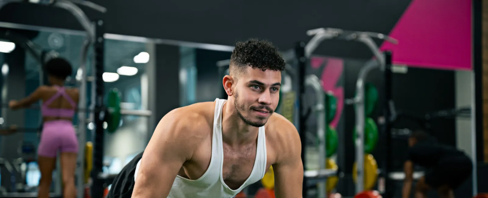 Man in white tank top doing push-ups on gym turf with weightlifting equipment and people in background