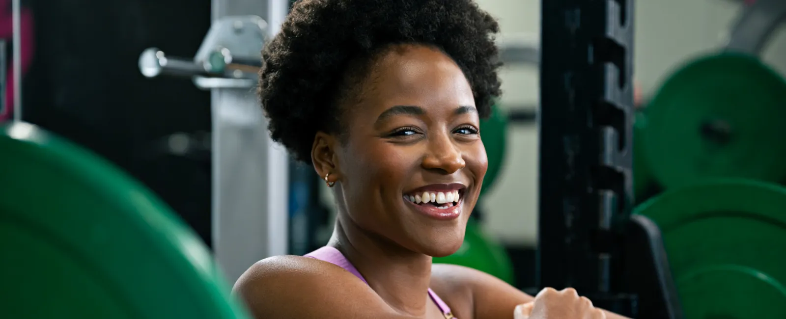 Smiling young woman resting on a barbell in a gym with weight plates in the background, ready for weightlifting.