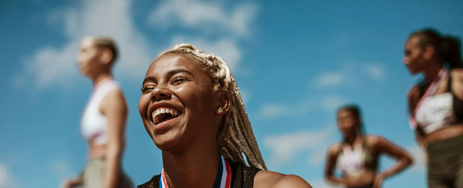 Smiling female athlete with medal around neck celebrating victory with fellow runners in background.