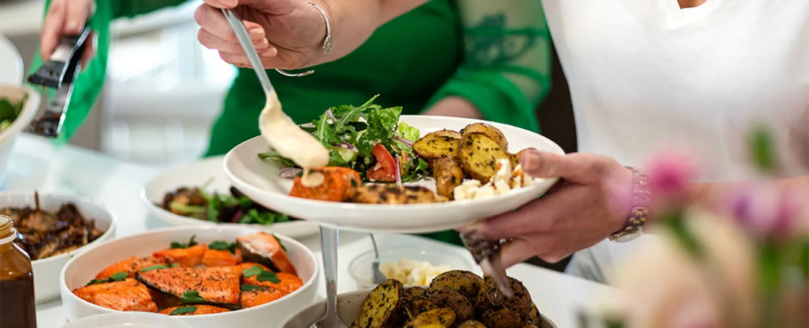 Person serving roasted potatoes and salad with grilled salmon at a buffet-style meal gathering.