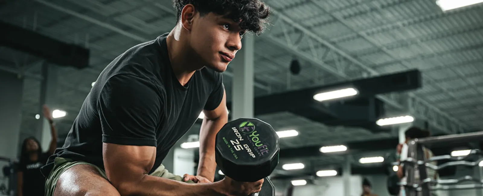 Young man with curly hair lifting a 25-pound dumbbell in a modern gym setting focused on strength training.