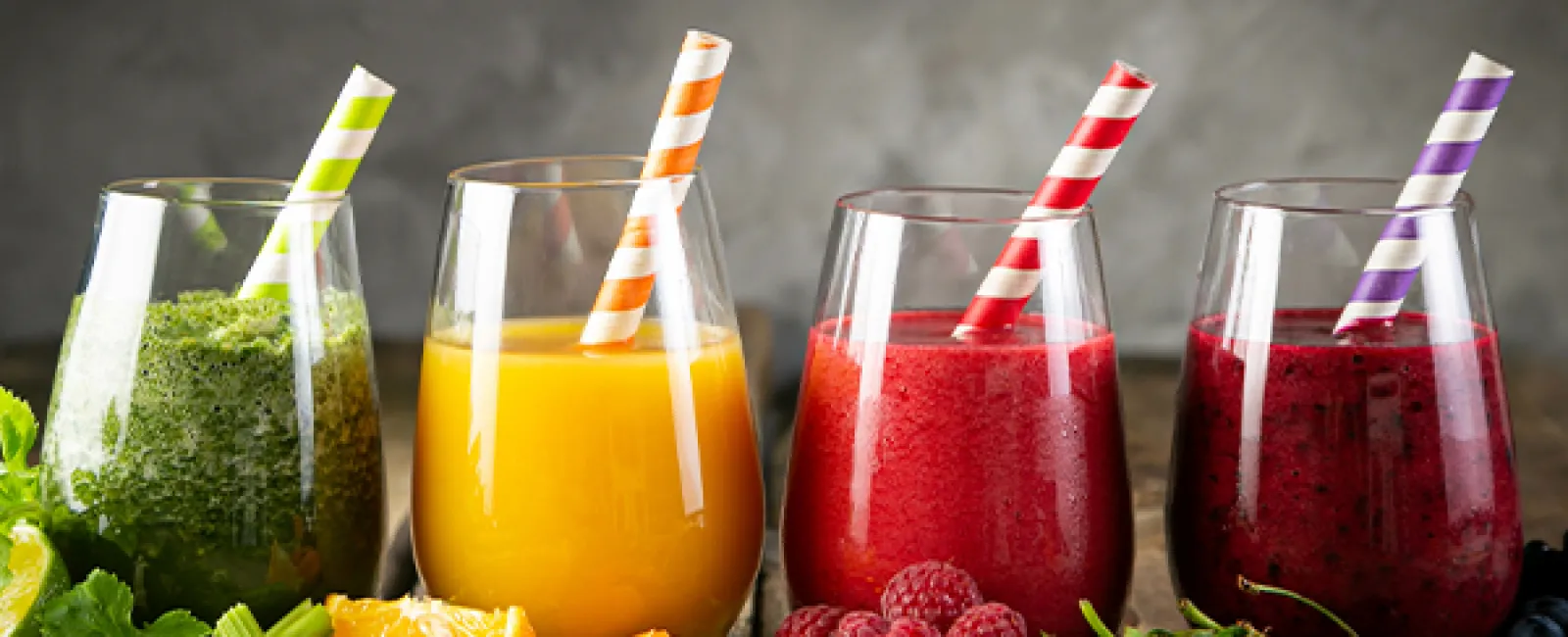Four glasses of green, orange, red, and purple smoothies with fresh fruits and vegetables on a wooden table.