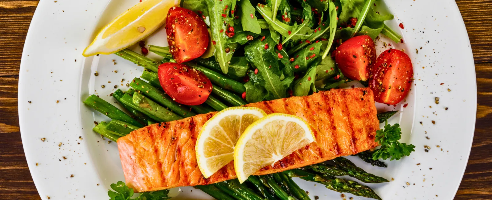 Grilled salmon served with lemon slices, asparagus, cherry tomatoes, and arugula salad on a white plate.