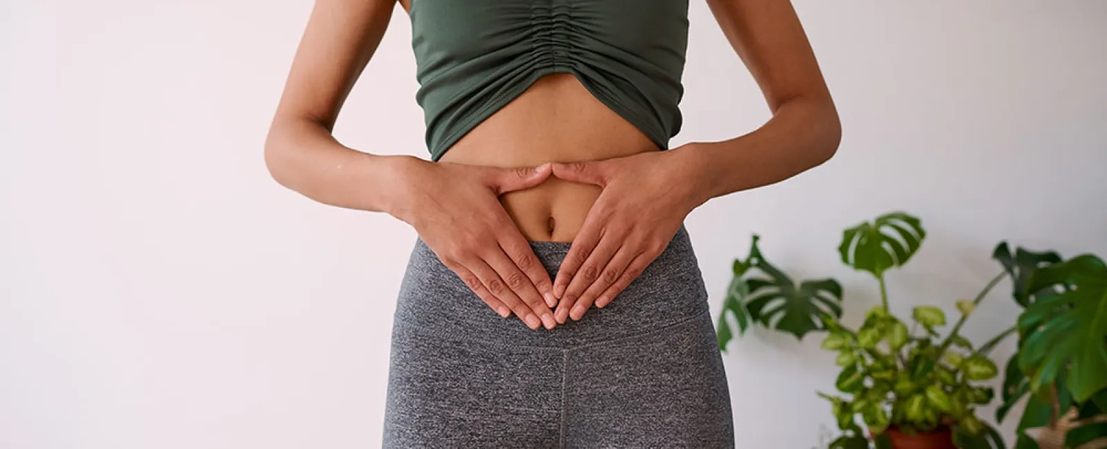 Woman in green crop top and gray leggings holding hands over stomach with houseplants in background