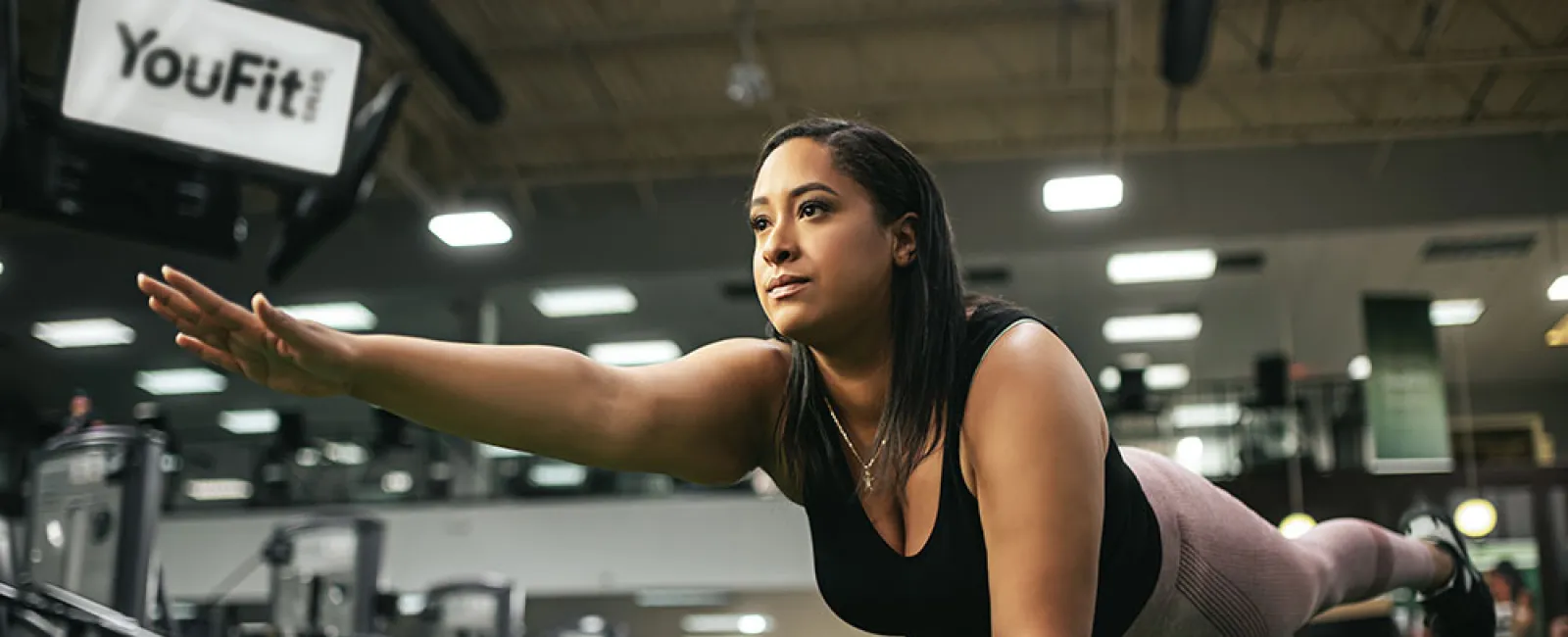 Woman performing bird dog exercise on turf at a modern gym with YouFit sign in the background
