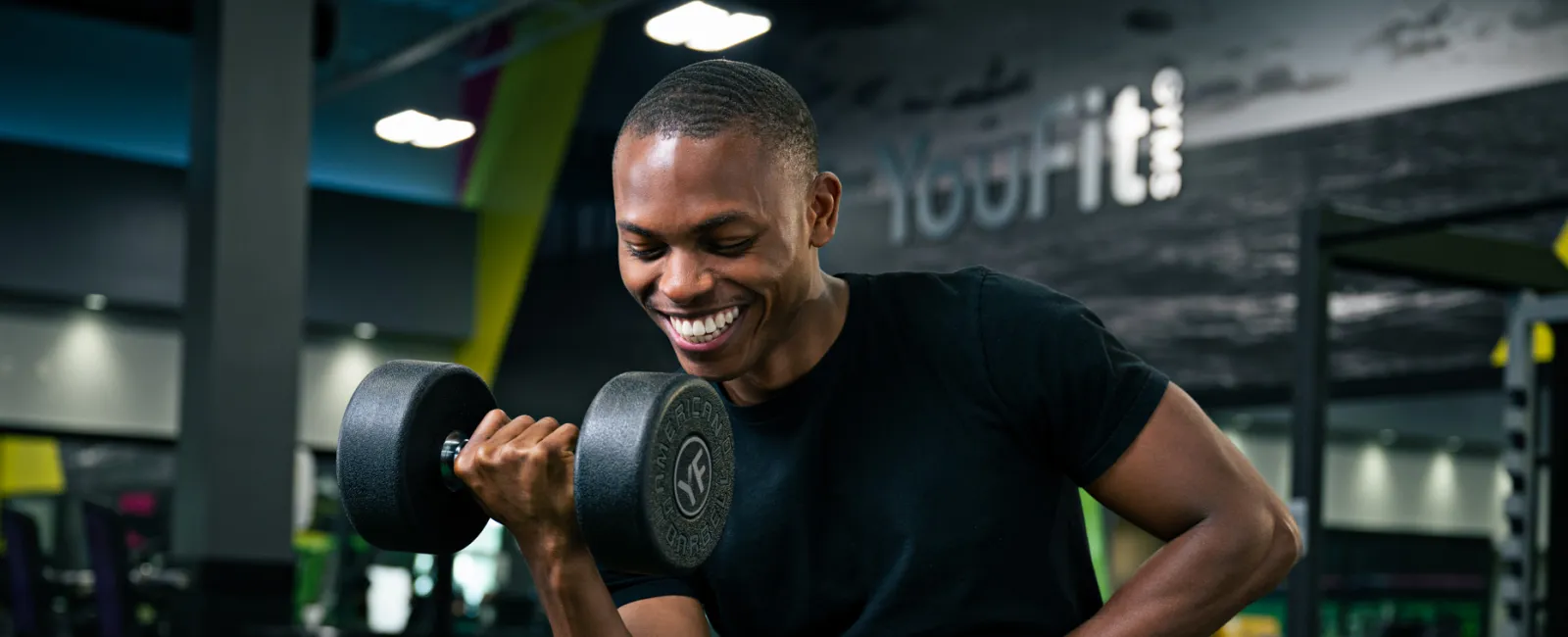Smiling young man lifting a dumbbell using a preacher curl bench in a modern gym setting.