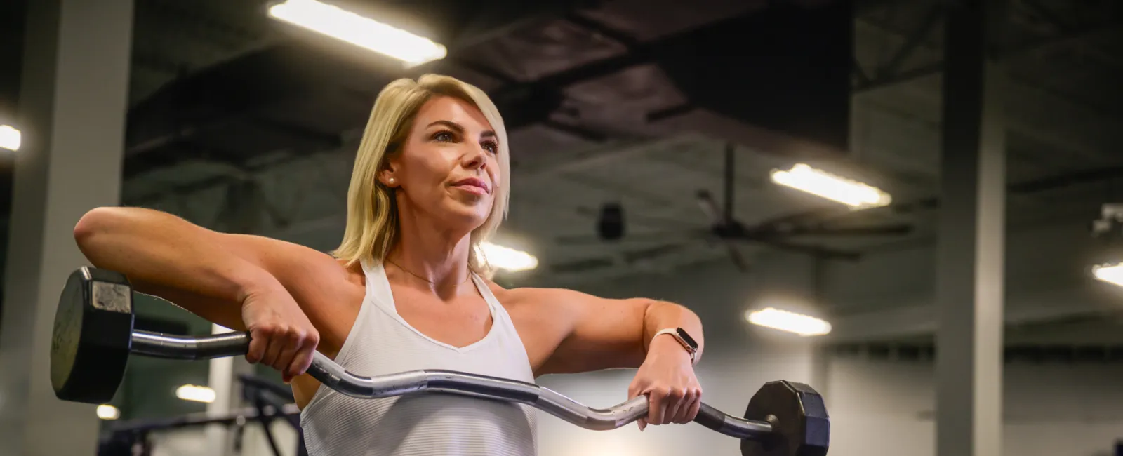 Woman in white tank top lifting barbell with weights in gym, focusing on strength training.