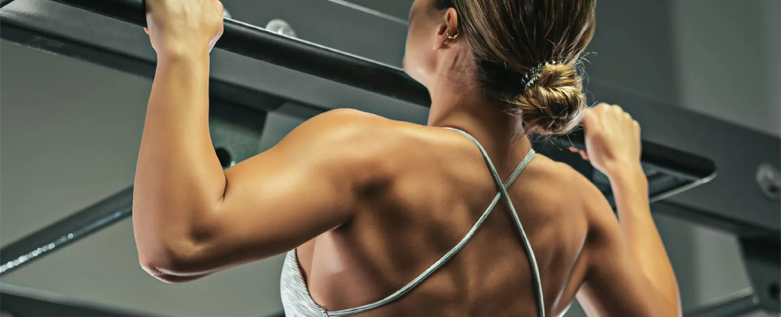 Woman performing pull-up exercise showcasing toned back muscles in gym setting with workout equipment.