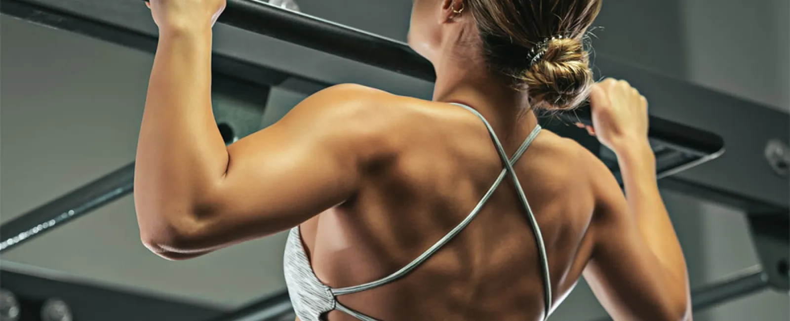 Woman performing pull-up exercise showcasing toned back muscles in gym setting with workout equipment.