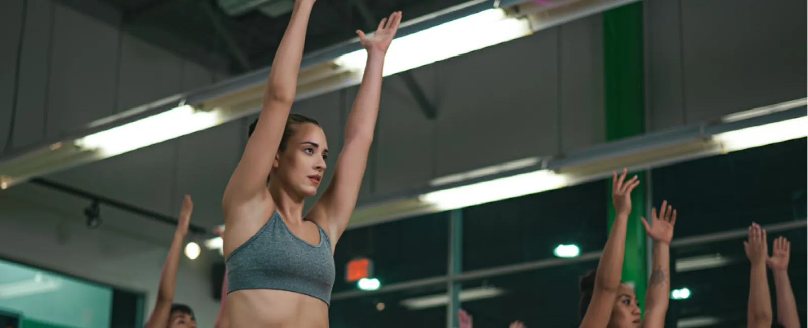Group of women in workout clothes doing yoga stretches with arms raised in a modern gym studio.