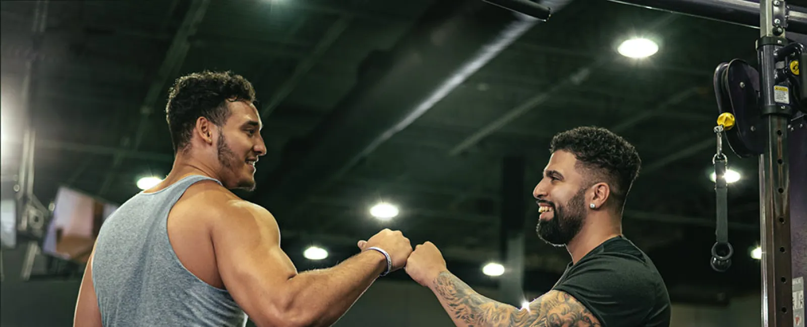 Two muscular men fist bump in a gym setting, celebrating a workout success with equipment in the background.