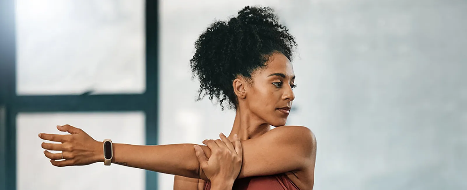 Woman in rust sportswear stretching her arm indoors with natural light from window.