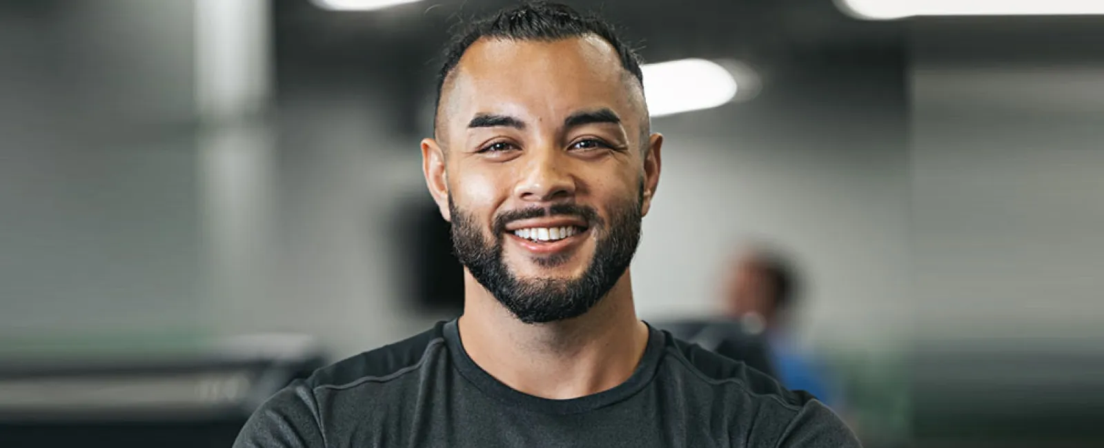 Smiling man with beard wearing a YouFit Gyms shirt and a tattooed arm in a gym setting.