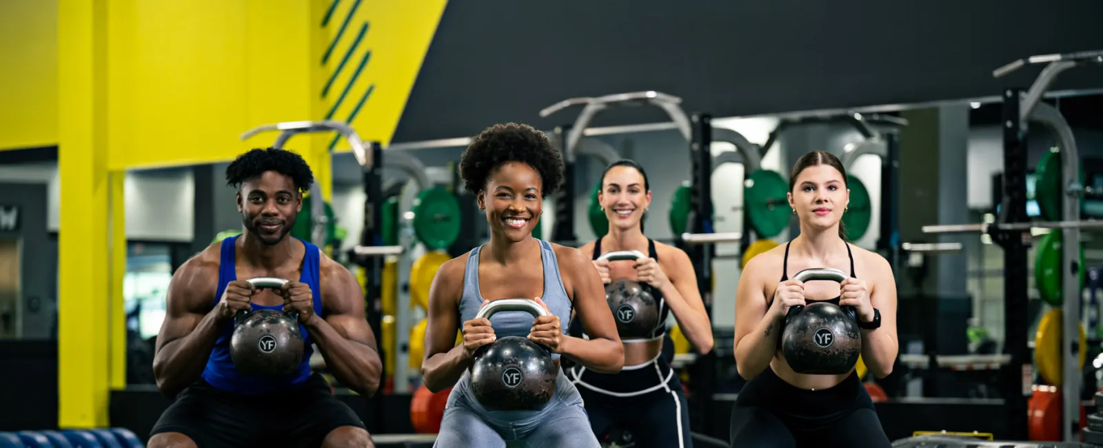 Four diverse people performing kettlebell squats together in a brightly lit modern gym with weights in the background