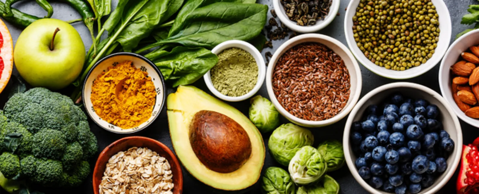 Fresh healthy superfoods including avocado, spinach, blueberries, broccoli, oats, nuts, and spices on a gray surface.