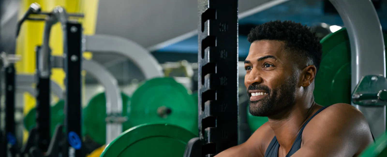 Smiling man resting on squat rack barbell with green weight plates in a colorful gym setting