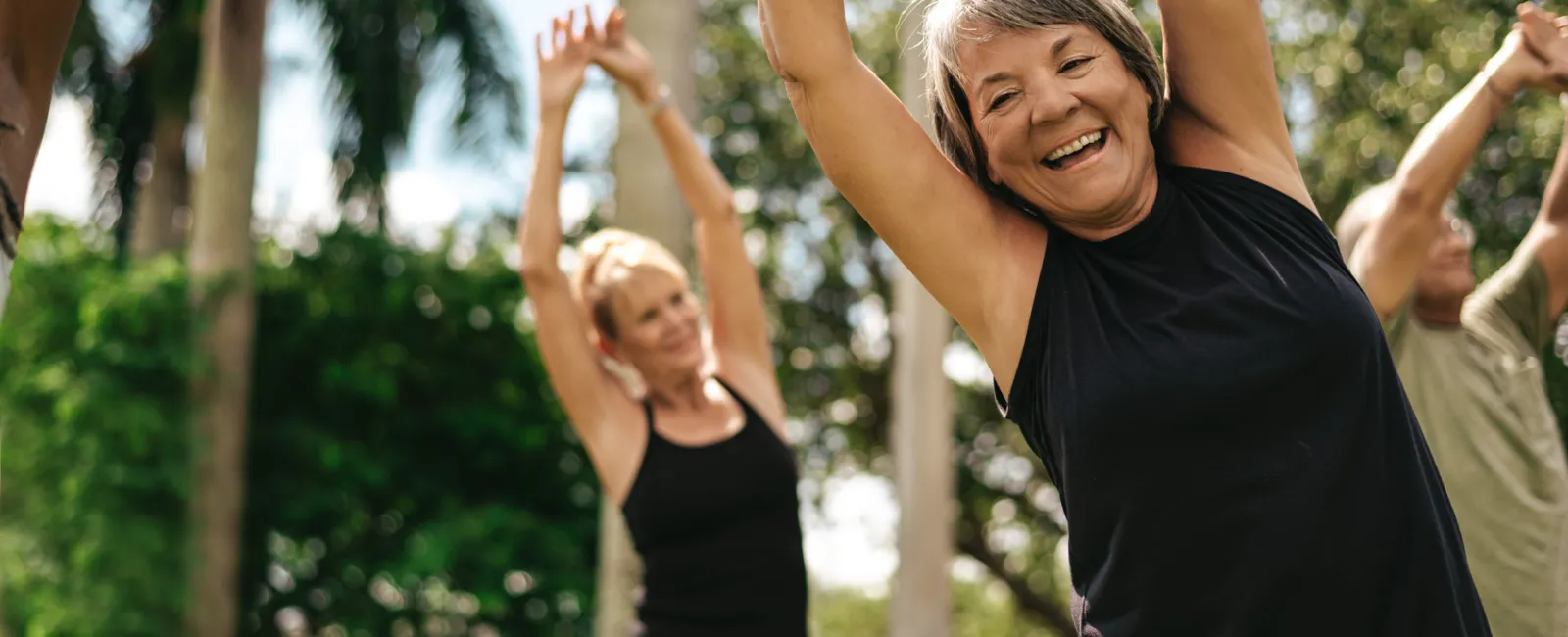 Smiling mature woman stretching arms up during outdoor group fitness class with palm trees in background
