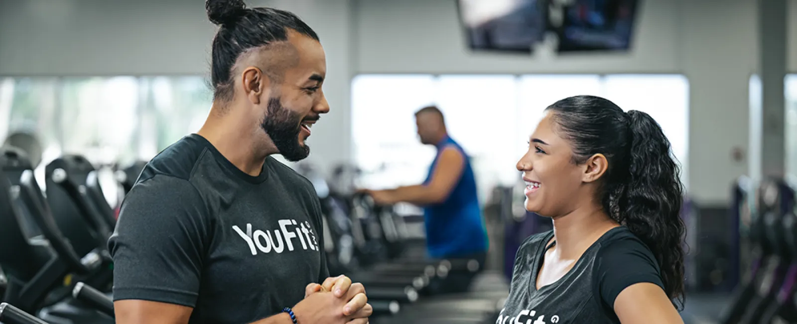 Male and female trainers wearing YouFit shirts talk and smile by gym treadmills in a fitness center.