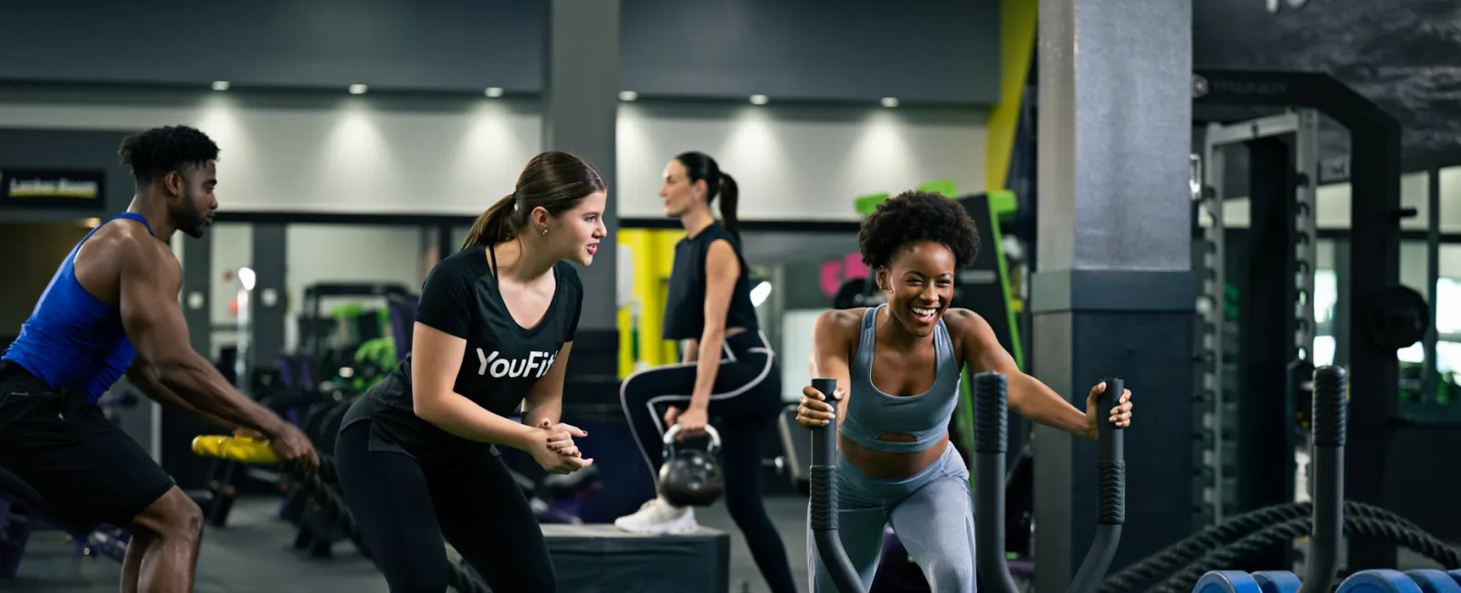 Group of diverse people exercising with battle ropes, sled push, and kettlebells in modern gym setting.