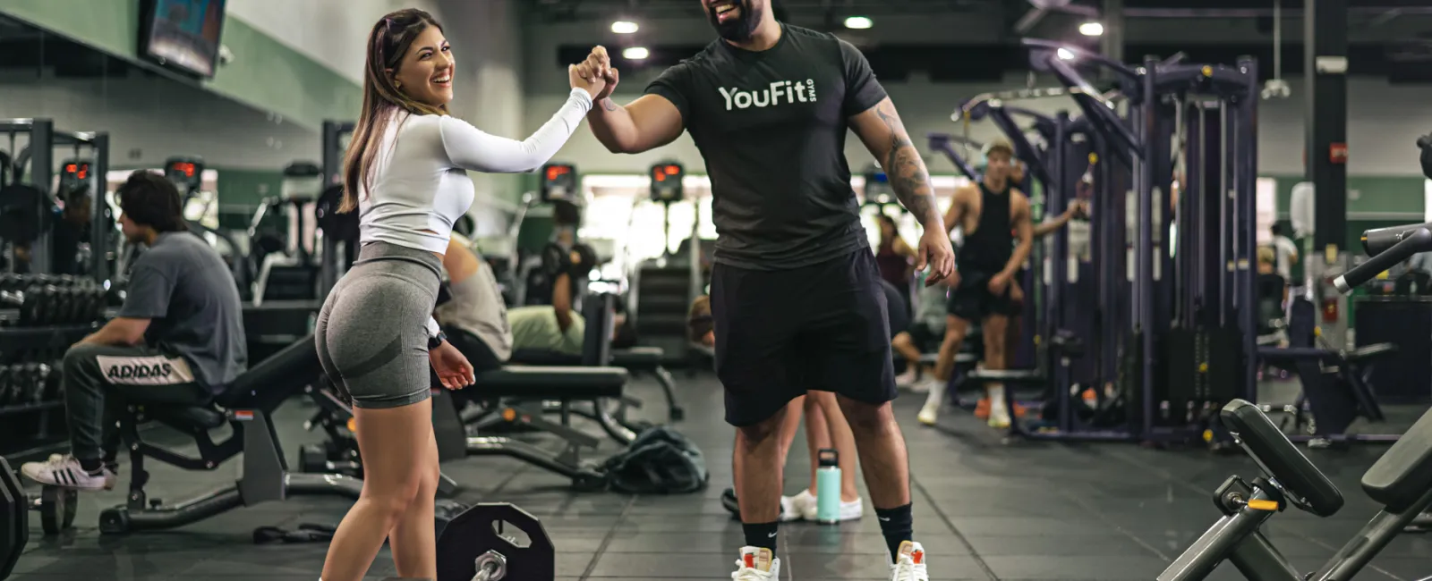 Two gym-goers fist bump in a busy fitness gym with weights and exercise equipment around.
