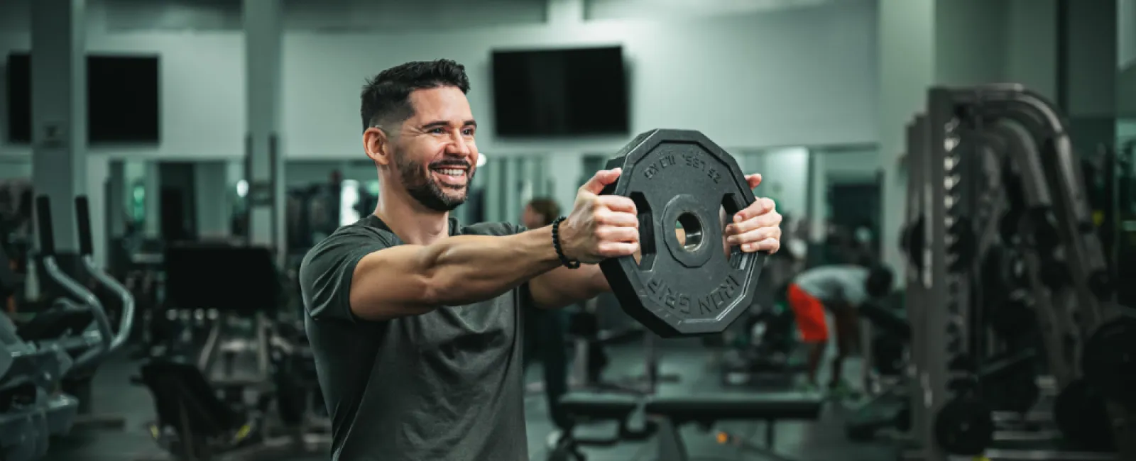 Smiling man lifting a weight plate in a modern gym with various exercise equipment in the background