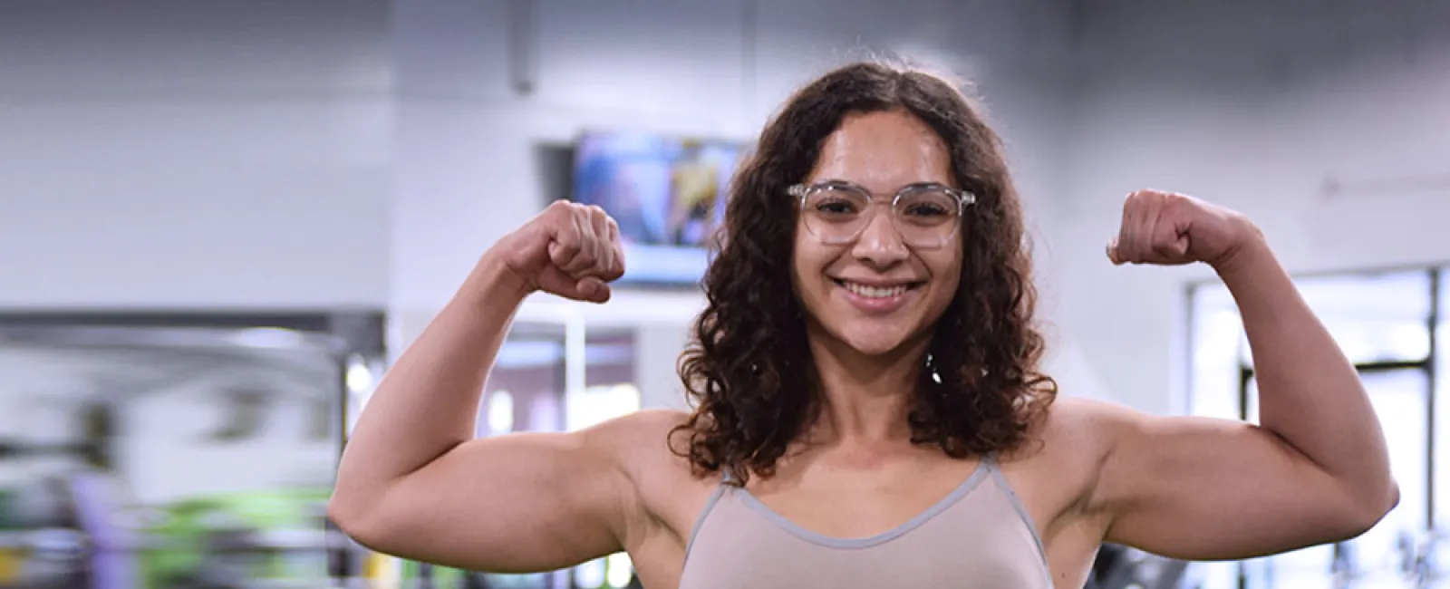 Smiling woman in gym flexing biceps wearing glasses and workout clothes showing fitness strength.