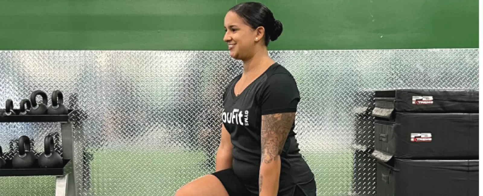 Woman performing a weighted lunge exercise holding dumbbells in a gym with green walls and artificial turf.
