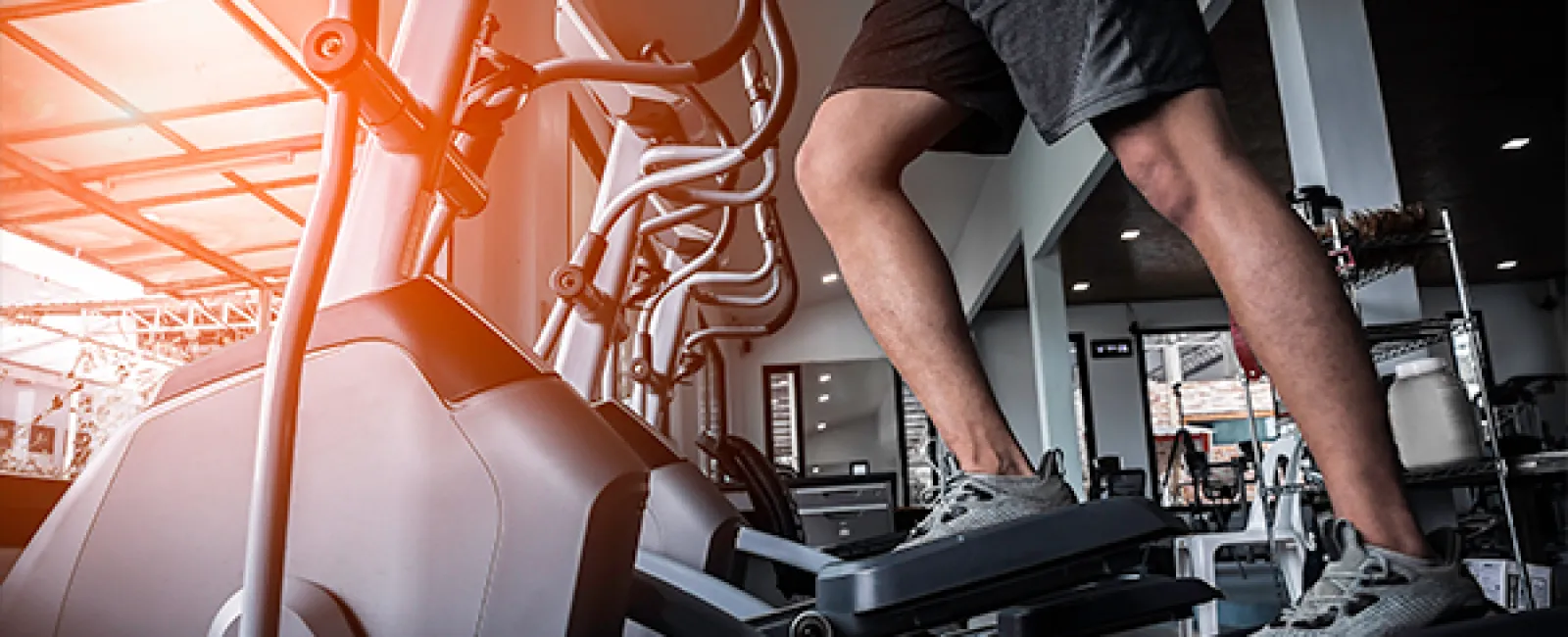 Person exercising on elliptical machine in gym with sunlight streaming through large windows.