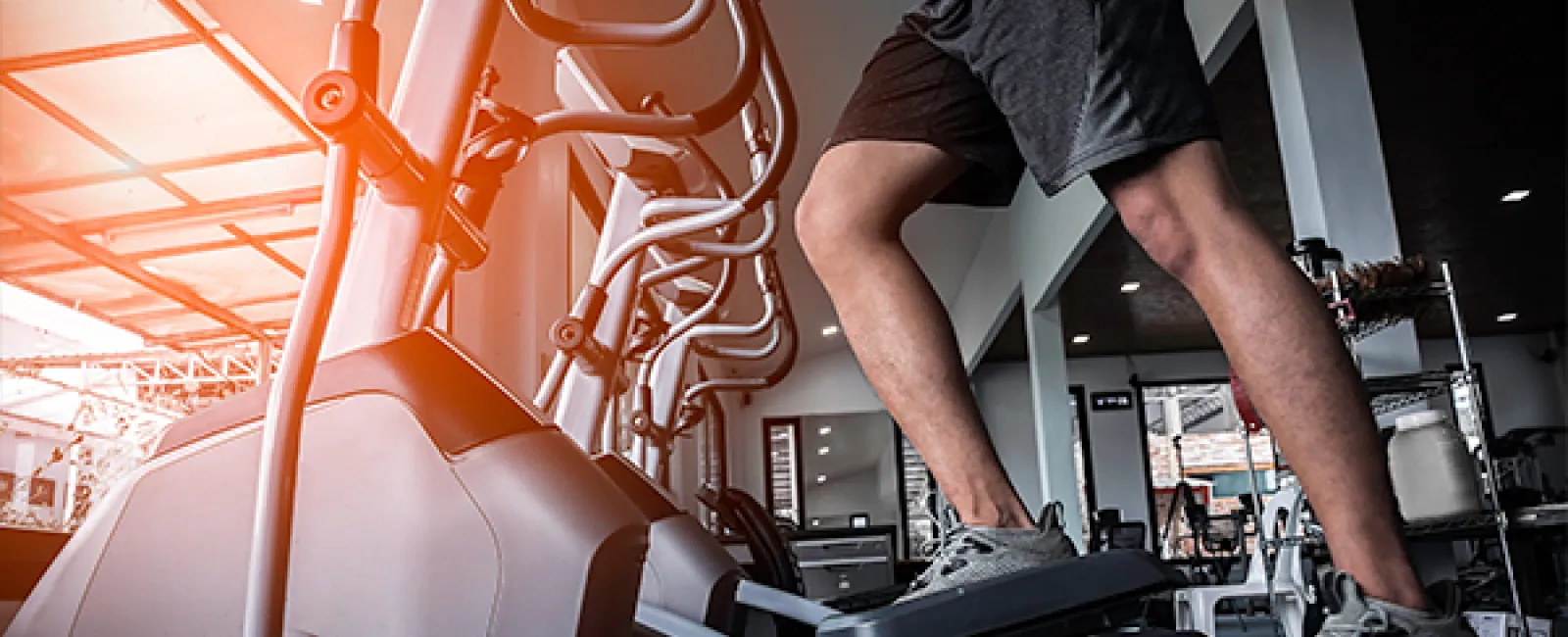 Person exercising on elliptical machine in gym with sunlight streaming through large windows.