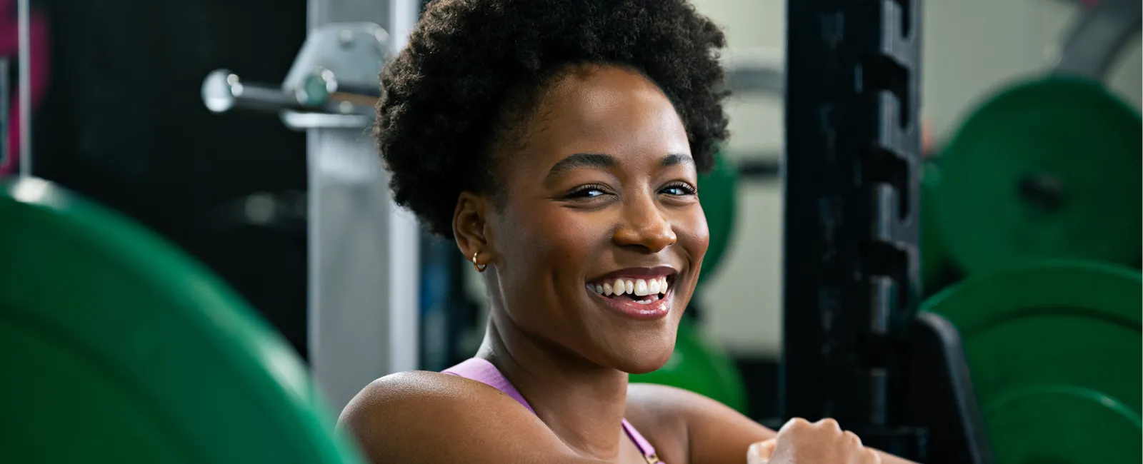 Smiling woman resting on a barbell rack in a gym surrounded by weight plates and fitness equipment.