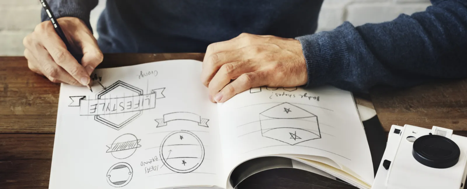Designer sketching logo concepts on a notebook at a wooden desk with a vintage camera nearby