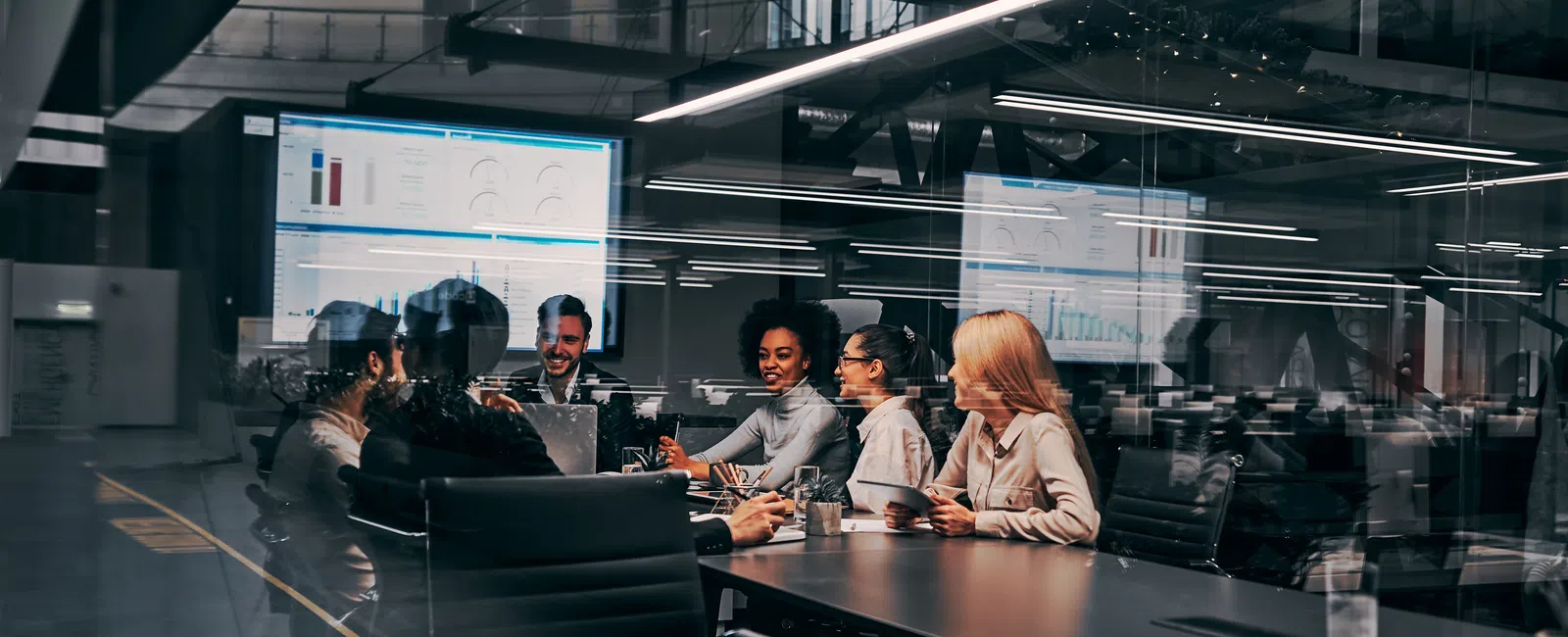 Diverse team meeting in modern glass office with digital charts on screen discussing business strategy.