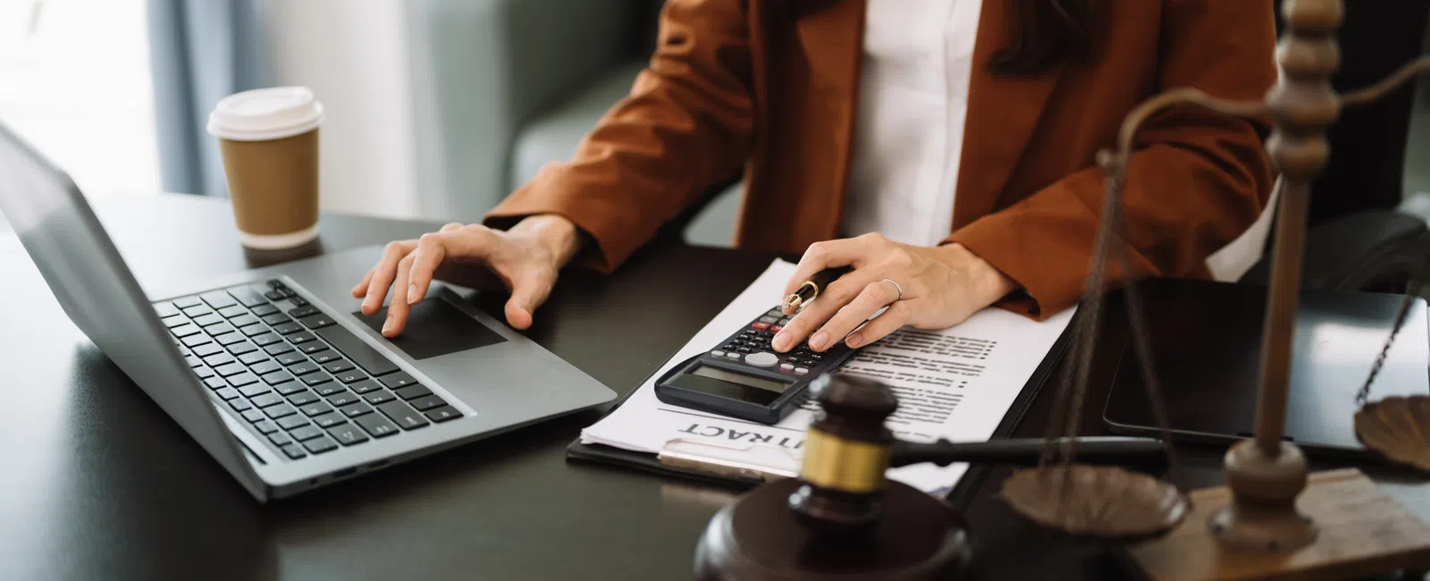 Legal professional working on laptop with contract documents, calculator, gavel, and scales of justice on desk.