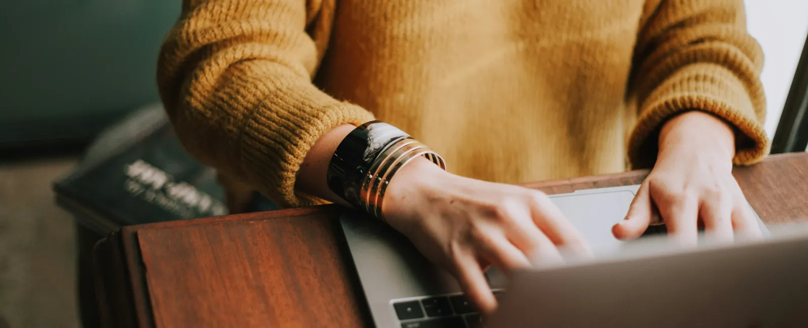 Person in mustard sweater typing on a laptop at a wooden desk with bracelets on the wrist.