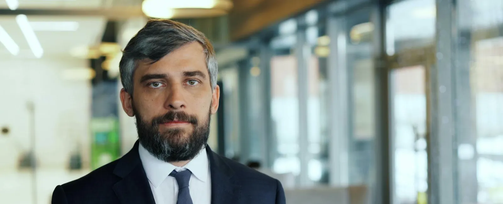 Confident businessman with gray beard wearing a dark suit and tie in a modern office setting.