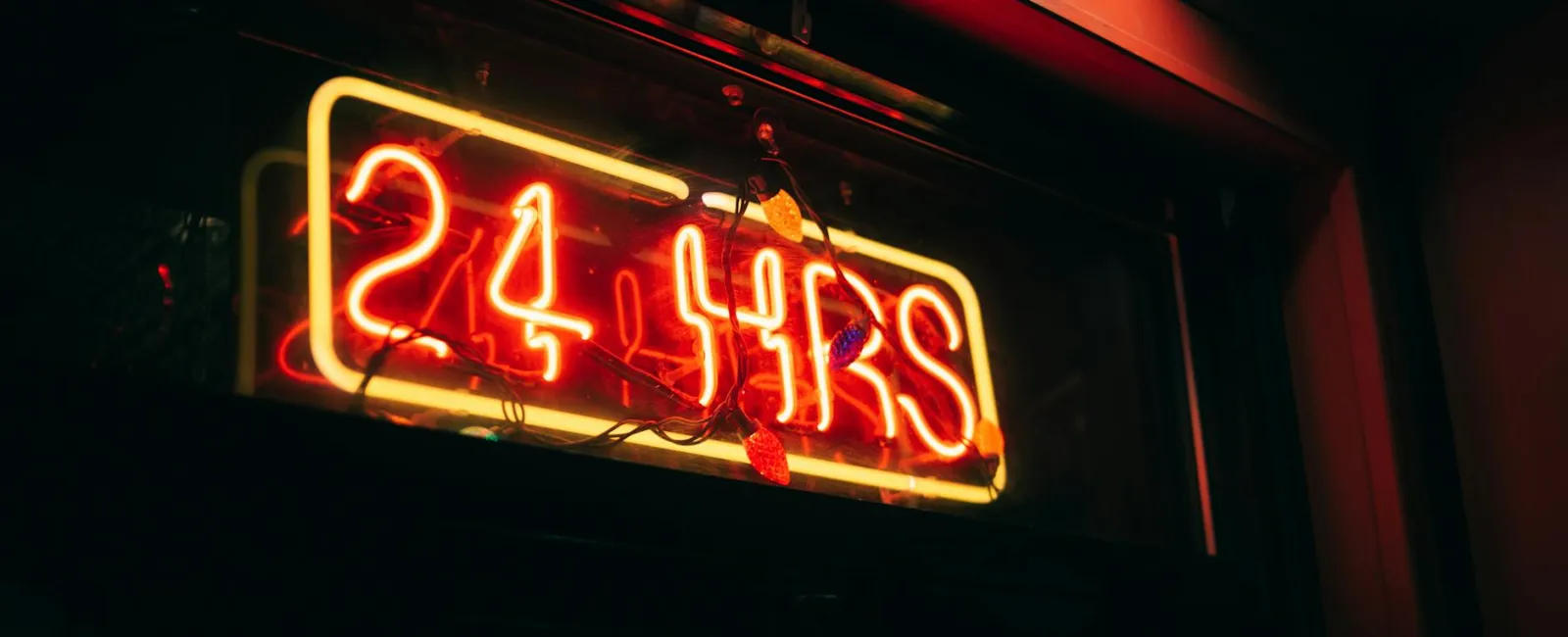 Red and yellow neon sign displaying 24 HRS inside a dimly lit building window with decorative lights.
