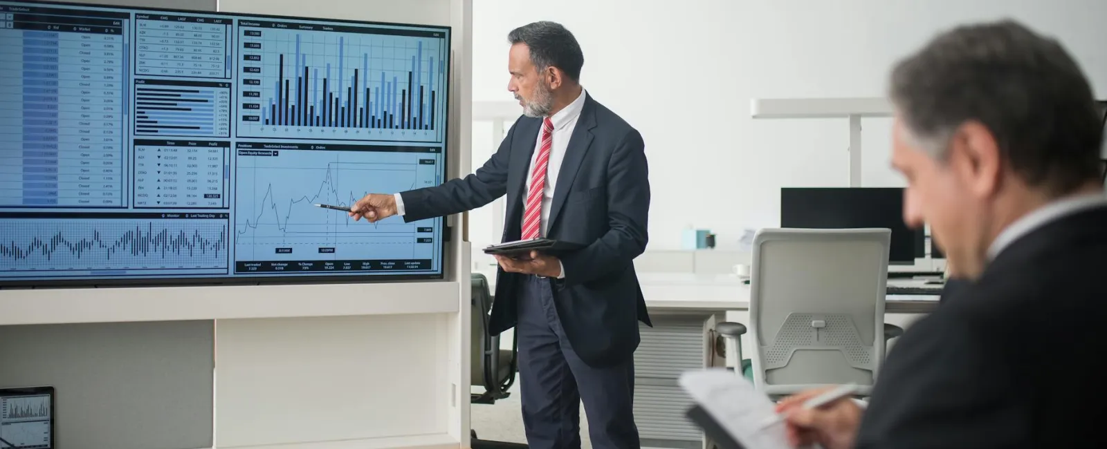 Businessman presenting financial charts and graphs on large screen while colleague takes notes in office.