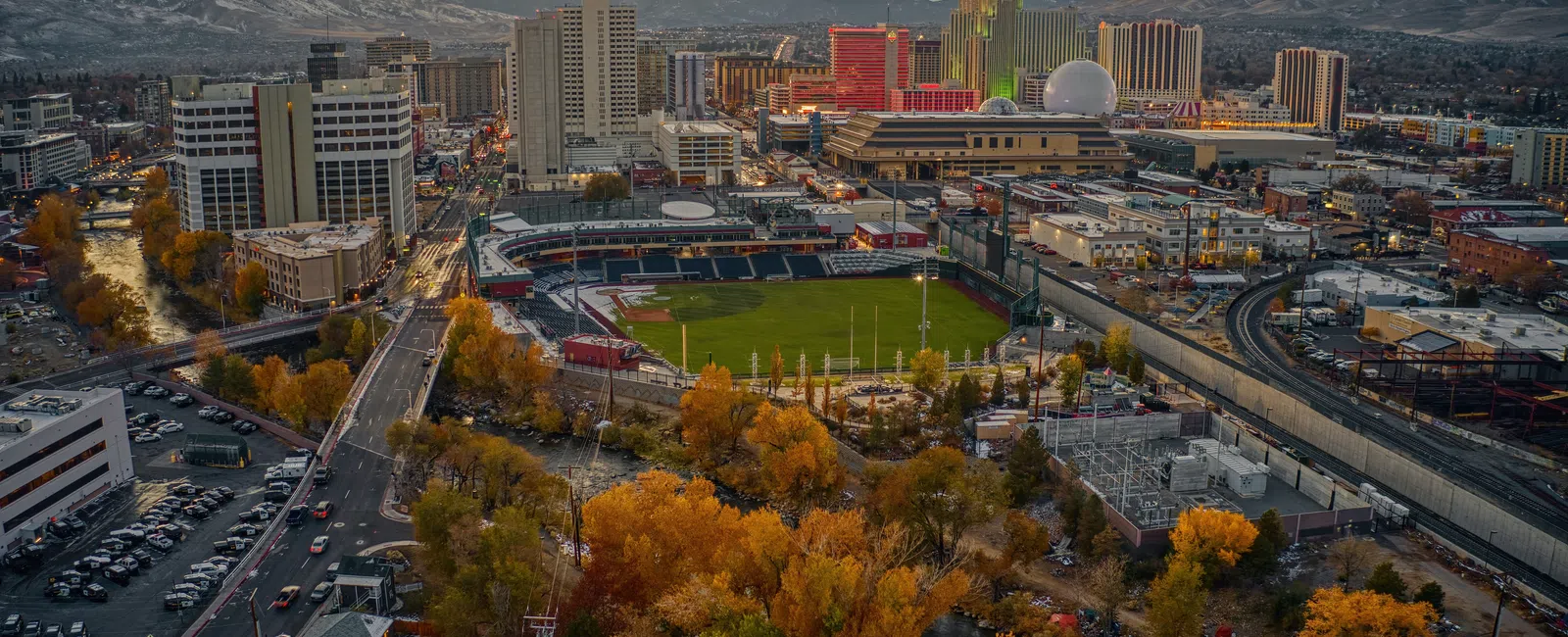 Aerial view of a city at sunset featuring a stadium, autumn trees, and distant mountains under a colorful sky.
