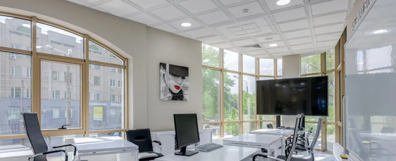 Bright modern office with white desks, black chairs, computers, large windows, and a wall-mounted TV screen.
