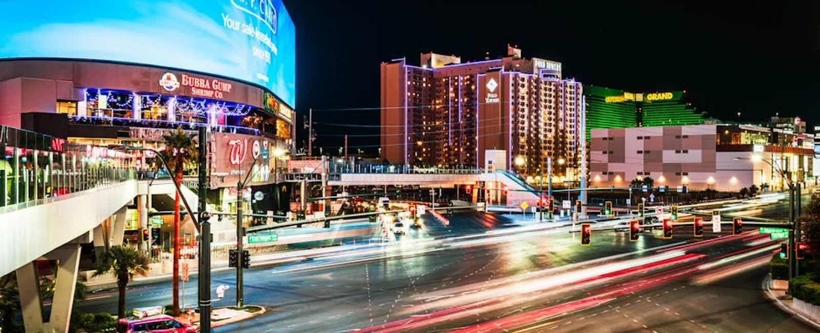Nighttime Las Vegas street scene with bright lights, casinos, traffic light trails, and tall buildings in vibrant colors.