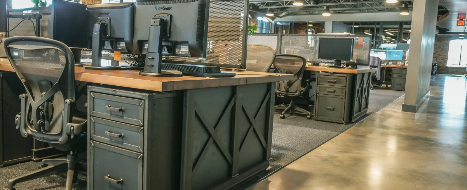 Modern open-plan office with wooden desks, ergonomic chairs, dual monitors, and industrial design elements.