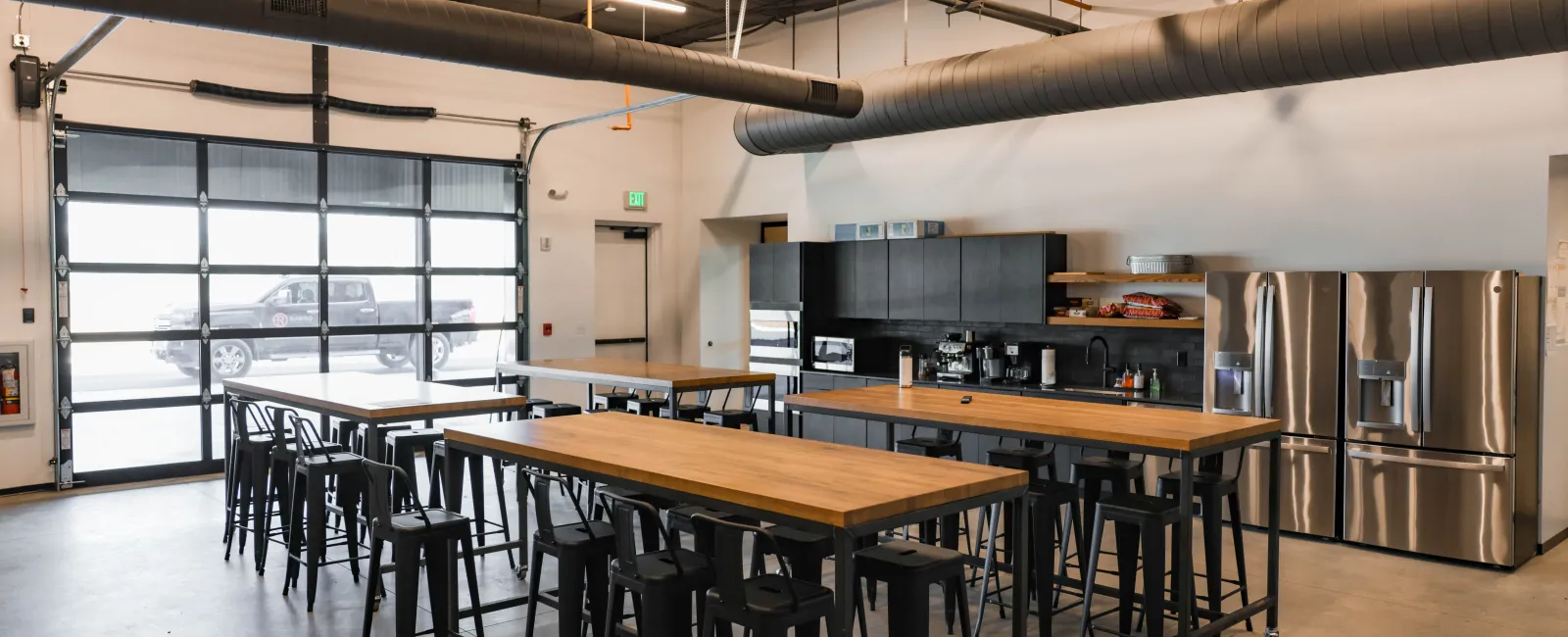 Modern industrial kitchen and dining area with wooden tables, black stools, stainless steel appliances, and exposed ceiling pipes