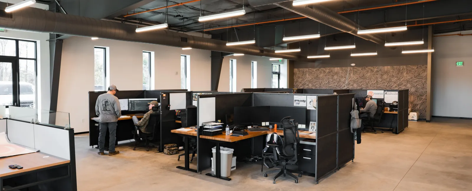 Modern office space with partitioned desks, computers, chairs, and two people working near windows.