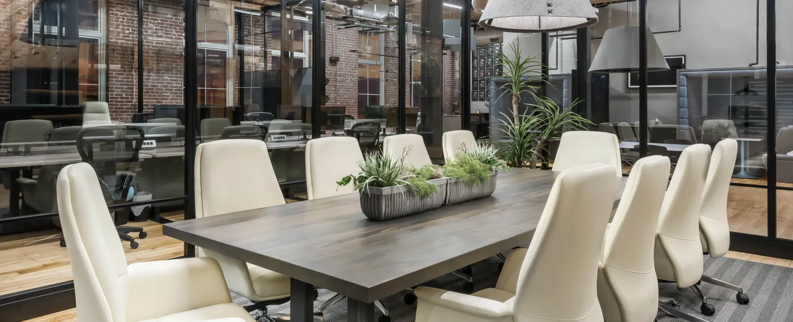 Modern conference room with a long wooden table, cream leather chairs, large pendant lights, and glass walls.