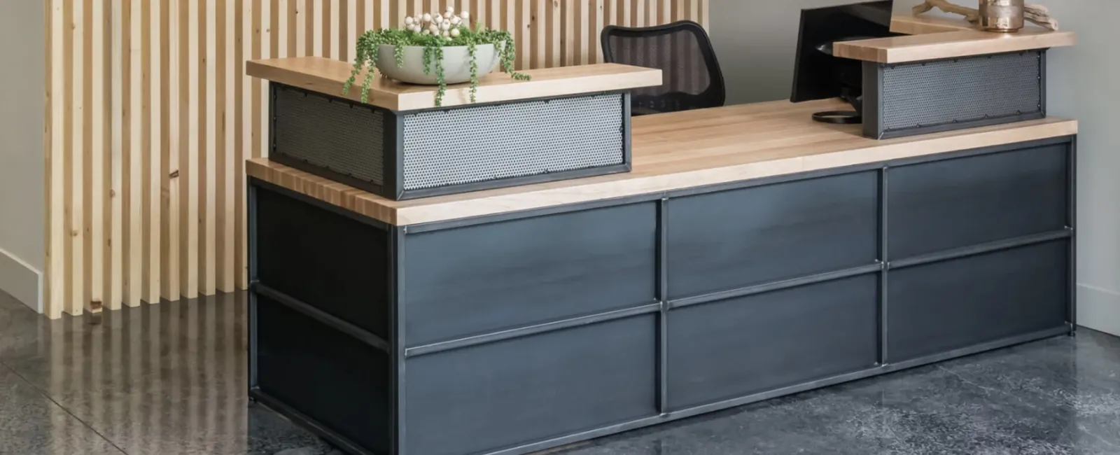 Modern reception desk with black metal base, wooden countertop, potted plants, and vertical wood paneling background.