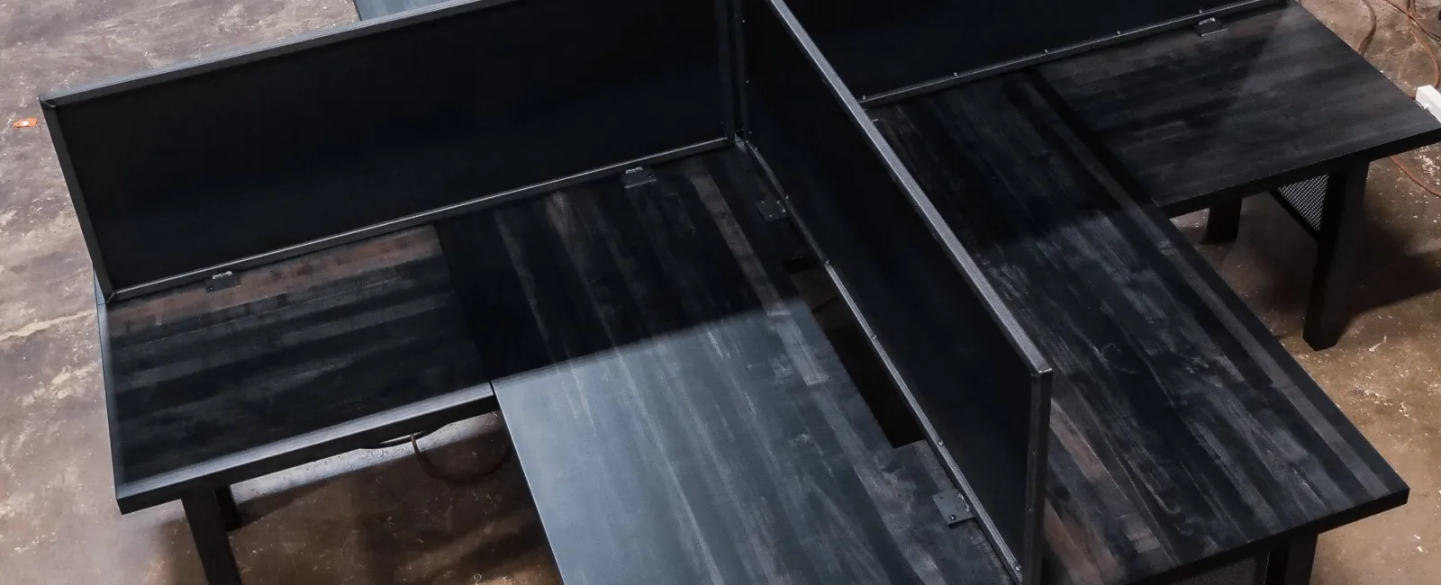 Overhead view of a modern black office workstation cluster with four desks and privacy panels in a workshop.